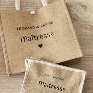 May include: A tan jute tote bag and a matching smaller pouch, both featuring white trim and black text that reads LE GRAND BAZAR DE Ma&icirc;tresse and LE PETIT BAZAR DE Ma&icirc;tresse with a small black heart icon below each phrase. The tote has a white button closure.