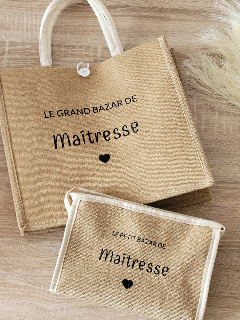 May include: A tan jute tote bag and a matching smaller pouch, both featuring white trim and black text that reads LE GRAND BAZAR DE Ma&icirc;tresse and LE PETIT BAZAR DE Ma&icirc;tresse with a small black heart icon below each phrase. The tote has a white button closure.