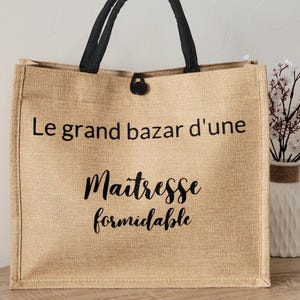 May include: A rectangular tan jute tote bag with black fabric handles and a black button closure. The front features black printed text that reads Le grand bazar d'une Maitresse formidable in a combination of sans-serif and script fonts.