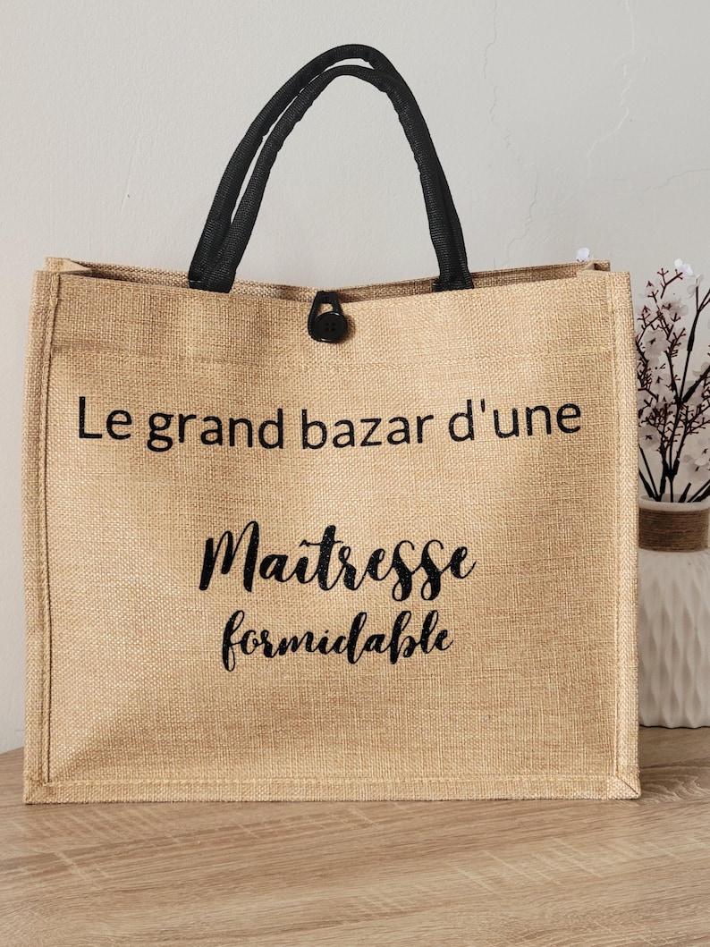 May include: A rectangular tan jute tote bag with black fabric handles and a black button closure. The front features black printed text that reads Le grand bazar d'une Maitresse formidable in a combination of sans-serif and script fonts.