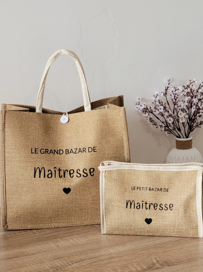 May include: A tan jute tote bag and a matching rectangular pouch, both featuring white trim and black text that reads LE GRAND BAZAR DE Ma&icirc;tresse and LE PETIT BAZAR DE Ma&icirc;tresse, each accented with a small black heart icon and a white button closure.