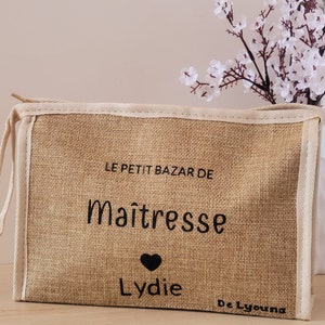 May include: A rectangular beige burlap pouch with a zipper closure and cream-colored trim. The front features black printed text that reads LE PETIT BAZAR DE Ma&icirc;tresse, a small black heart icon, the name Lydie, and the text De Lyouna in the corner.