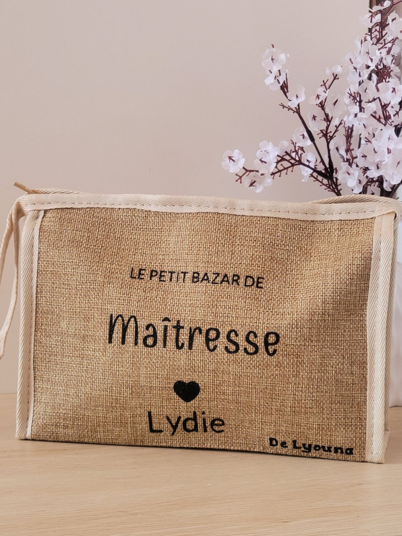 May include: A rectangular beige burlap pouch with a zipper closure and cream-colored trim. The front features black printed text that reads LE PETIT BAZAR DE Ma&icirc;tresse, a small black heart icon, the name Lydie, and the text De Lyouna in the corner.