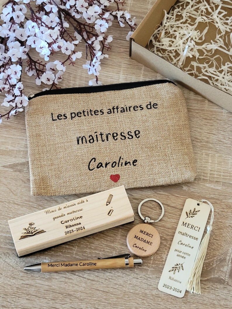 May include: A set of teacher appreciation gifts including a burlap pencil pouch with the text "Les petites affaires de maitresse Caroline", a wooden bookmark with the text "MERCI maitresse Caroline pour cette ann&eacute;e Rihanna 2023-2024", a wooden keychain with the text "MERCI MADAME Caroline", a wooden eraser with the text "Merci de m'avoir aid&eacute; &agrave; grandir ma&icirc;tresse Caroline Rihanna 2023-2024", and a wooden pen with the text "Merci Madame Caroline".