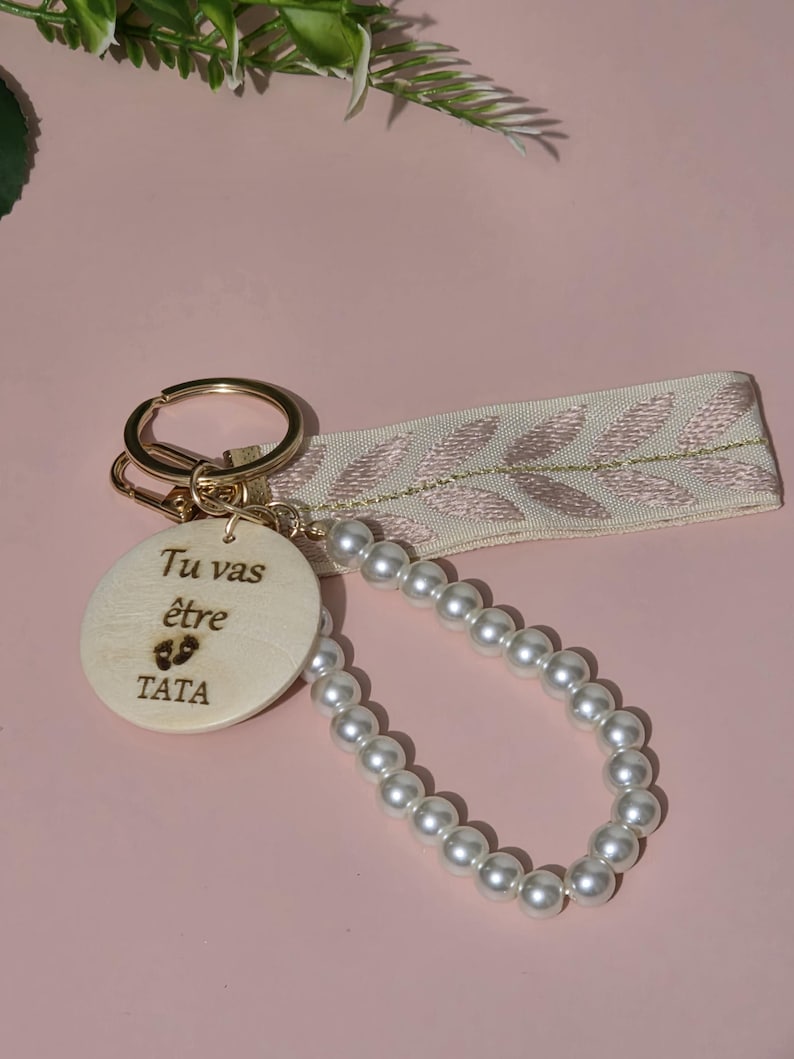 K&ouml;nnte beinhalten: Ein Holzschl&uuml;sselanh&auml;nger mit einer Perlenkette. Der Holzanh&auml;nger hat den Text "Tu vas &ecirc;tre TATA" und ein kleines Fu&szlig;abdruck-Symbol.