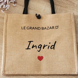 May include: A rectangular tan jute tote bag with black fabric handles and a black button closure. The front features the black text LE GRAND BAZAR D' and the name Ingrid in a script font, followed by a small red heart graphic centered below.