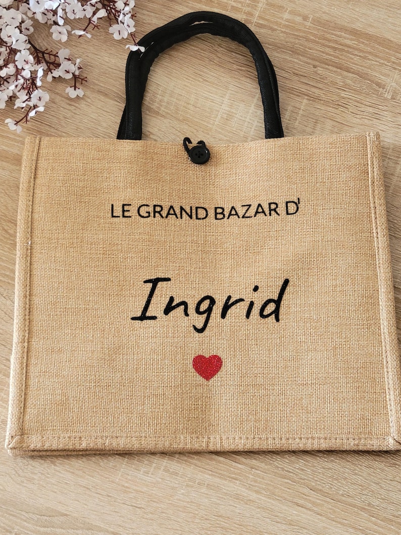May include: A rectangular tan jute tote bag with black fabric handles and a black button closure. The front features the black text LE GRAND BAZAR D' and the name Ingrid in a script font, followed by a small red heart graphic centered below.