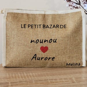 May include: A rectangular beige burlap pouch with a zipper closure and white fabric trim. The front features black text reading LE PETIT BAZARDE, nounou, and Aurore, with a small red glitter heart between the last two lines and M&eacute;lina in the corner.