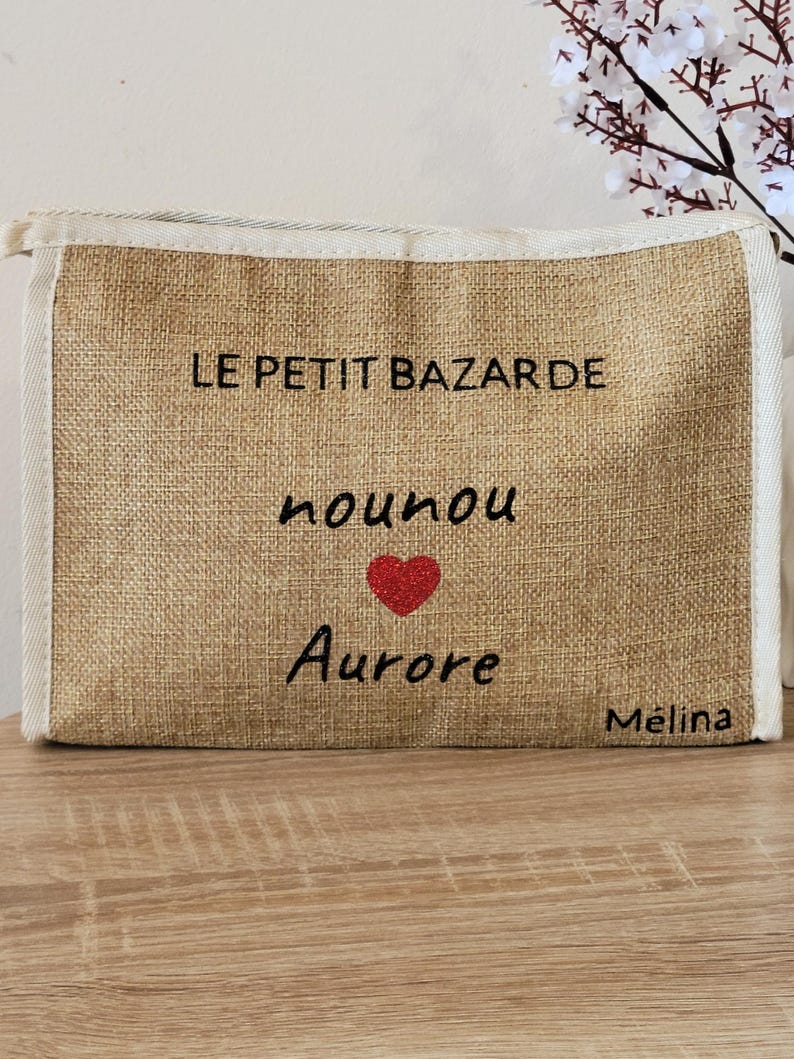 May include: A rectangular beige burlap pouch with a zipper closure and white fabric trim. The front features black text reading LE PETIT BAZARDE, nounou, and Aurore, with a small red glitter heart between the last two lines and M&eacute;lina in the corner.