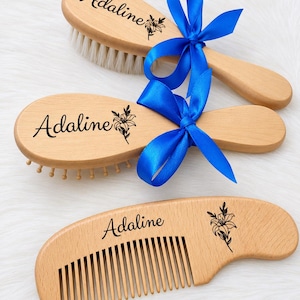 May include: Two wooden hairbrushes and one wooden comb, each engraved with the name Adaline and a floral illustration. The two brushes feature soft bristles and rounded wooden pins respectively, and both are adorned with a bright blue satin ribbon.