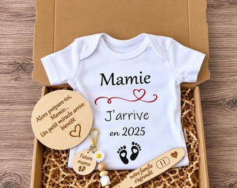 May include: A white baby bodysuit with black text reading Mamie J'arrive en 2025 and a red heart graphic. Included are a round wooden disc with French text, a keychain with a daisy and wooden beads, and a wooden stick with the text notre famille s'agrandit.