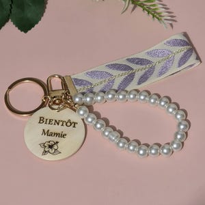 K&ouml;nnte beinhalten: Ein Holzschl&uuml;sselanh&auml;nger mit einer Perlenkette und einem hellvioletten Band mit einem Blattmuster. Der Holzanh&auml;nger tr&auml;gt den Text "BIENT&Ocirc;T Mamie" und ein kleines Blumenmuster.