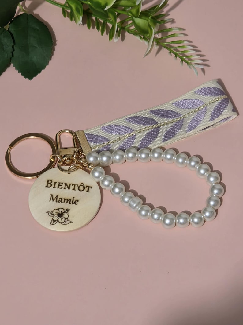 K&ouml;nnte beinhalten: Ein Holzschl&uuml;sselanh&auml;nger mit einer Perlenkette und einem hellvioletten Band mit einem Blattmuster. Der Holzanh&auml;nger tr&auml;gt den Text "BIENT&Ocirc;T Mamie" und ein kleines Blumenmuster.