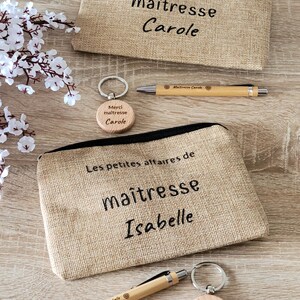 May include: Two brown burlap zippered pouches with the text "Les petites affaires de maitresse" and the names "Carole" and "Isabelle" embroidered on them. Each pouch is paired with a wooden keychain with the text "Merci maitresse" and the name "Carole" or "Isabelle" engraved on it. Each pouch also comes with a wooden pen with the text "Maitresse Carole" or "Maitresse Isabelle" engraved on it.