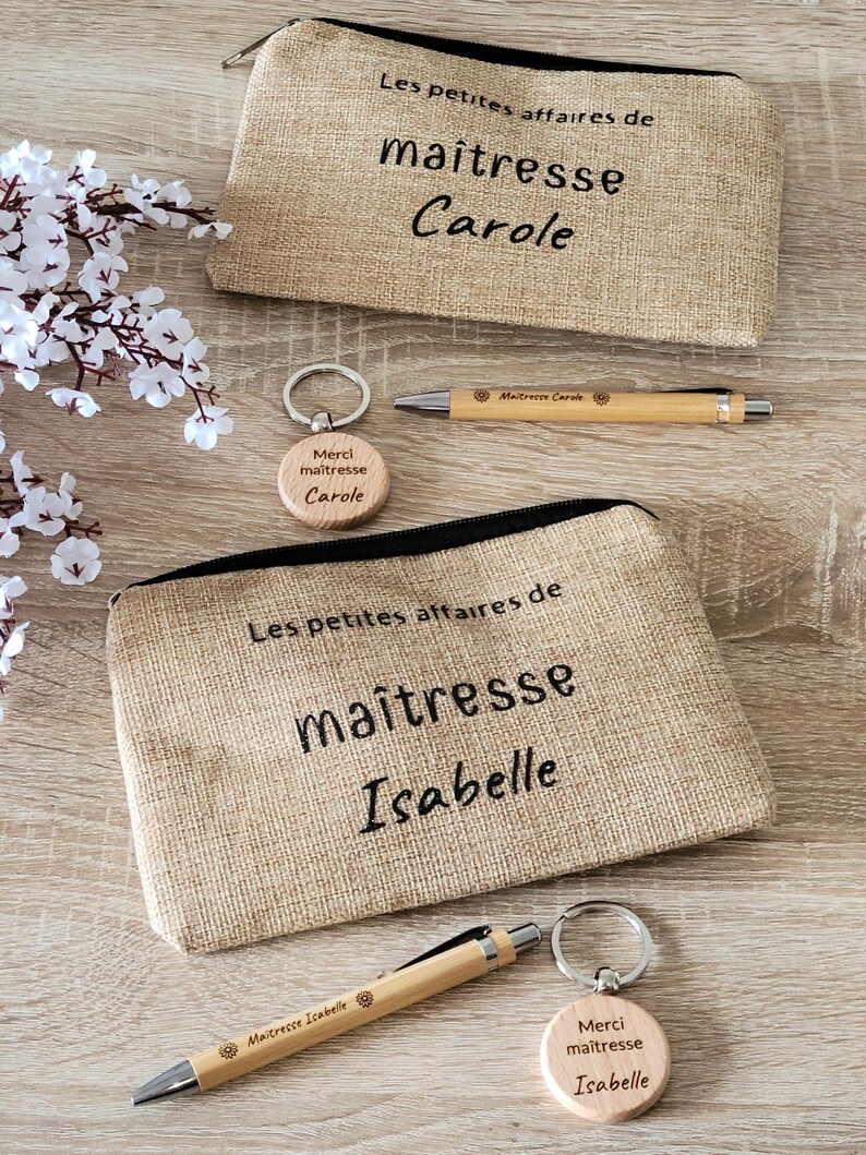 May include: Two brown burlap zippered pouches with the text "Les petites affaires de maitresse" and the names "Carole" and "Isabelle" embroidered on them. Each pouch is paired with a wooden keychain with the text "Merci maitresse" and the name "Carole" or "Isabelle" engraved on it. Each pouch also comes with a wooden pen with the text "Maitresse Carole" or "Maitresse Isabelle" engraved on it.