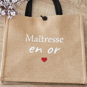 May include: A rectangular tan jute tote bag with two black fabric handles and a small black button closure. The front features white text that reads Ma&icirc;tresse en or with a small red heart graphic centered below the text on the woven fabric.