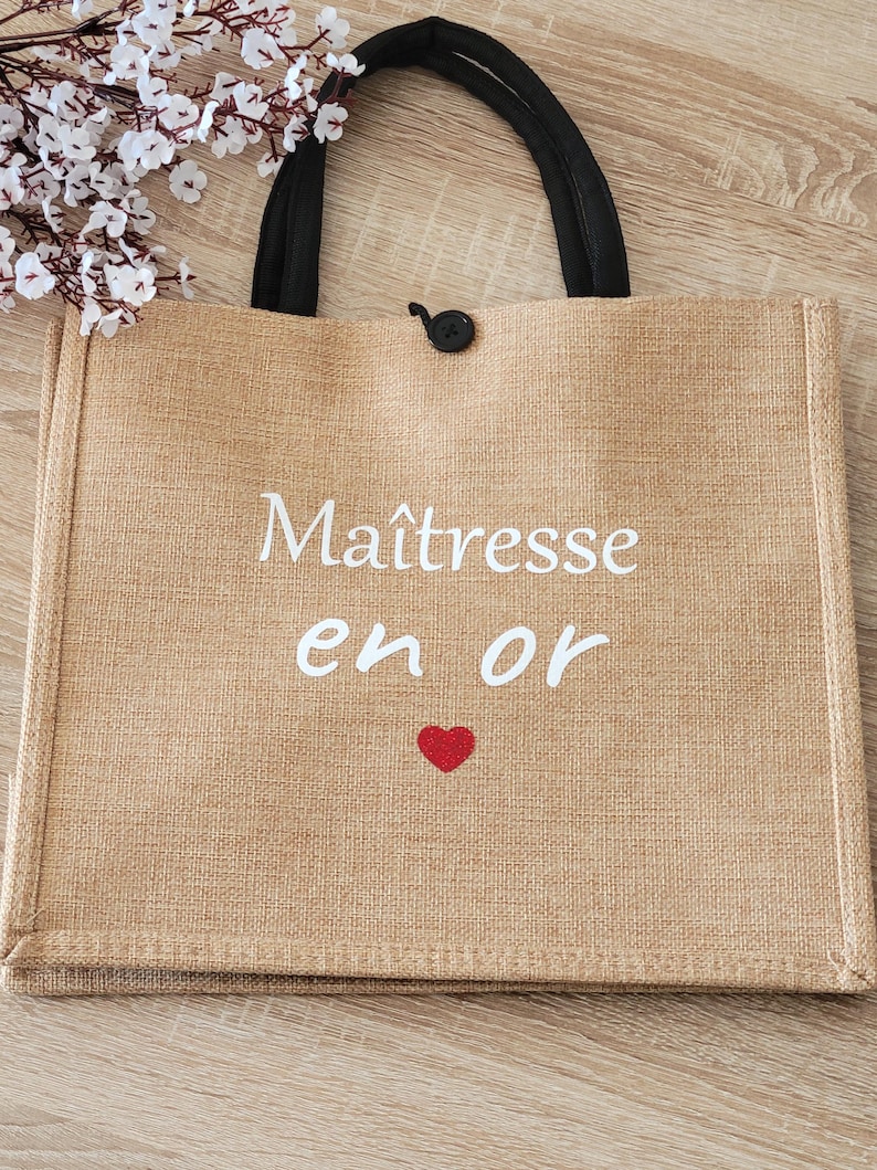 May include: A rectangular tan jute tote bag with two black fabric handles and a small black button closure. The front features white text that reads Ma&icirc;tresse en or with a small red heart graphic centered below the text on the woven fabric.