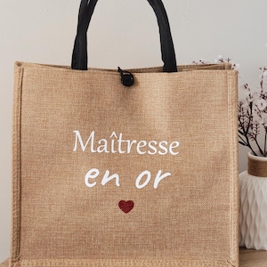 May include: A tan rectangular tote bag with black fabric handles and a black button closure. The front features the white text "Ma&icirc;tresse en or" above a small red glitter heart. The bag has a textured, woven appearance and a structured shape.