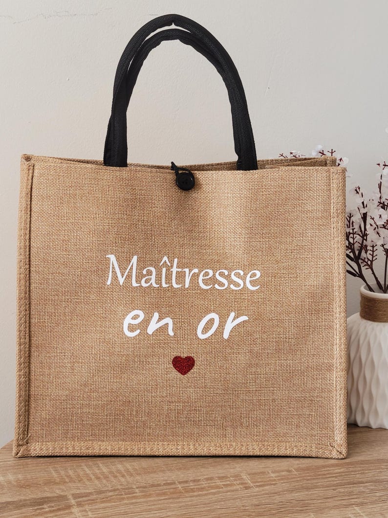 May include: A tan rectangular tote bag with black fabric handles and a black button closure. The front features the white text "Ma&icirc;tresse en or" above a small red glitter heart. The bag has a textured, woven appearance and a structured shape.
