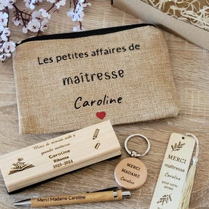 May include: A teacher appreciation gift set with a burlap makeup bag that says "Les petites affaires de maitresse Caroline", a wooden pencil holder with the text "Merci de m'avoir aid&eacute; &agrave; grandir maitresse Caroline Rihanna 2023-2024", a wooden keychain with the text "Merci Madame Caroline", a wooden bookmark with the text "Merci maitresse Caroline pour cette ann&eacute;e Rihanna 2023-2024", and a pen.