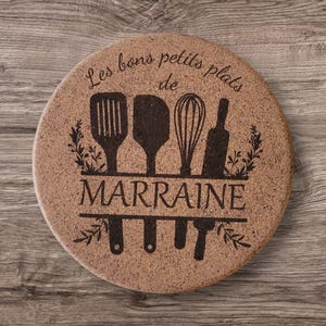 Peut inclure: Sous-plat rond en liège avec un motif d'ustensiles de cuisine, dont une spatule, une cuillère en bois, un fouet et un rouleau à pâtisserie. Le texte "Les bons petits plats de MARRAINE" est gravé sur le sous-plat.