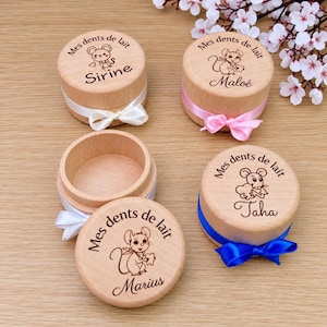 May include: Four round wooden boxes for baby teeth, each engraved with a mouse illustration and the text Mes dents de lait. The lids feature names Sirine, Maloé, Taha, and Marius, with white, pink, and blue satin ribbons tied around the bases.