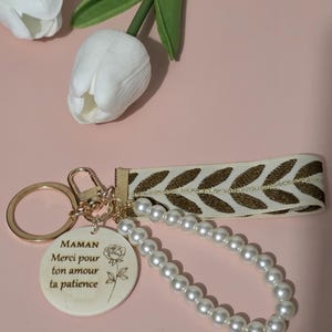 K&ouml;nnte beinhalten: Ein wei&szlig;er Keramik-Schl&uuml;sselanh&auml;nger mit einem goldenen Ring und einer Perlenkette. Der Schl&uuml;sselanh&auml;nger hat ein Rosenmotiv und den Text "MAMAN Merci pour ton amour ta patience".