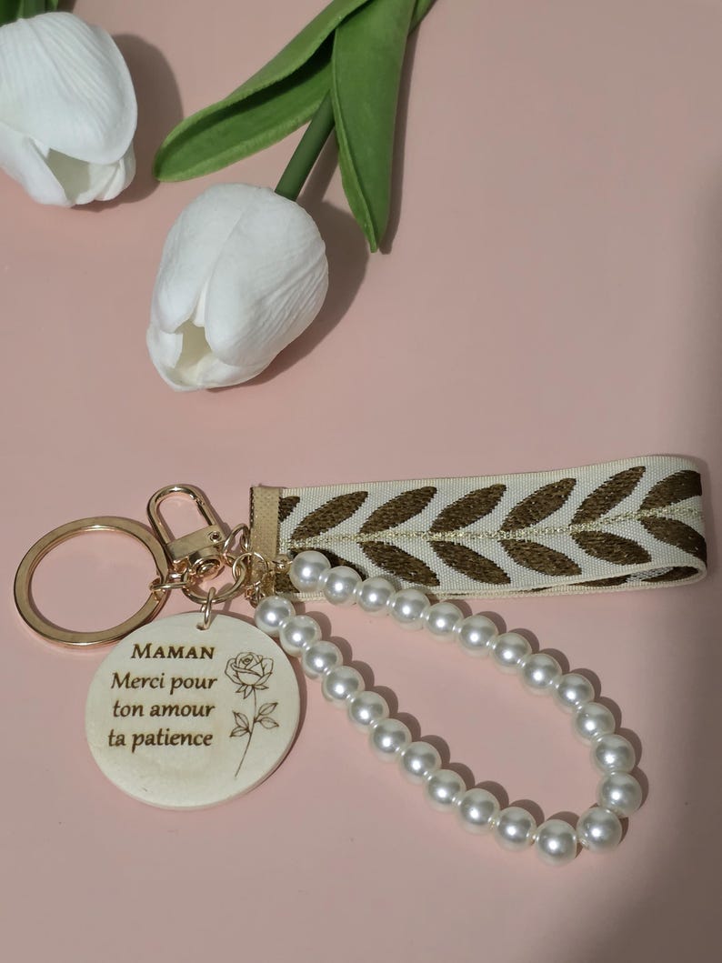 K&ouml;nnte beinhalten: Ein wei&szlig;er Keramik-Schl&uuml;sselanh&auml;nger mit einem goldenen Ring und einer Perlenkette. Der Schl&uuml;sselanh&auml;nger hat ein Rosenmotiv und den Text "MAMAN Merci pour ton amour ta patience".