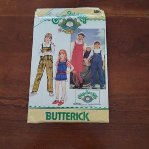 Puede incluir: Un sobre de patrón de costura Butterick para ropa de Cabbage Patch Kids, modelo 680. El frente presenta ilustraciones y fotos de niños vistiendo petos, un vestido de tirantes y pantalones cortos, con el texto Cabbage Patch Kids y Butterick.