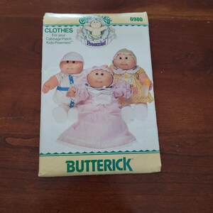 Op de afbeelding: Een Butterick naaipatroon-envelop, nummer 6980, voor Cabbage Patch Kids Preemie poppenkleding. De voorkant toont drie poppen in een witte outfit, een roze jurk met een witte kraag en een gele jurk met patroon en een mutsje.