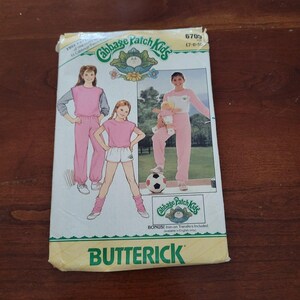 Puede incluir: Un sobre de patrón de costura Butterick para ropa de Cabbage Patch Kids, modelo 6705, tallas 7-8-10. El frente presenta ilustraciones y una fotografía de niños con ropa deportiva rosa y blanca, incluyendo una transferencia termoadhesiva extra.