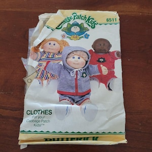 Puede incluir: Un sobre de patrón de costura Cabbage Patch Kids, número 6511, con ilustraciones de tres muñecos con varios atuendos, incluyendo una chaqueta gris con capucha y ribete rojo, una camisa roja y un vestido a rayas, con el texto CLOTHES For your Cabbage Patch Kids.