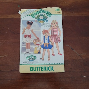 Puede incluir: Un sobre de patrón de costura Butterick Cabbage Patch Kids, número 3257, talla 5-6-6X. El frente presenta ilustraciones de niños con tres estilos de vestidos diferentes, incluyendo un vestido blanco con faja roja, uno azul y uno de rayas rojas.