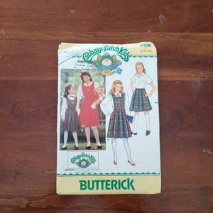 Puede incluir: Un sobre de patrón de costura Butterick para ropa de Cabbage Patch Kids, modelo 3356, tallas 7-8-10. El frente muestra ilustraciones de niños con un vestido rojo liso, un vestido a cuadros y una falda a cuadros con camisas blancas.