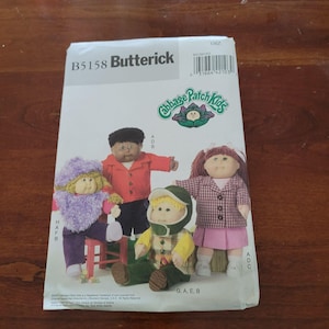 Op de afbeelding: Een Butterick B5158 naaipatroon-envelop voor Cabbage Patch Kids poppen. De voorzijde toont illustraties van vier poppen in verschillende outfits, waaronder een paars pluizig jack met capuchon, een rood overhemd, een groene pet en een geruit jasje.
