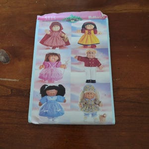 Puede incluir: Un sobre de patrón de costura Butterick 5115 Cabbage Patch Kids con seis ilustraciones de muñecas con varios atuendos, que incluyen un vestido rojo con capucha, un vestido amarillo, un vestido rosa, una chaqueta militar roja, un vestido azul y un vestido floral.
