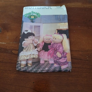 Könnte beinhalten: Ein Butterick-Schnittmusterumschlag für Cabbage Patch Kids-Puppen, Nummer 5206. Die Vorderseite zeigt drei Puppen mit Wollhaaren in goldfarbenen, rosafarbenen und violetten Kleidern vor einem Hintergrund mit einem karierten Boden.