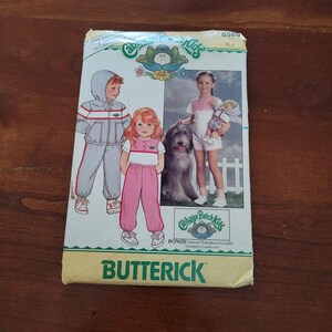 Può includere: Una busta di cartamodello Butterick per abbigliamento Cabbage Patch Kids, taglia 6X. Il fronte presenta illustrazioni e una fotografia di bambini che indossano una tuta con cappuccio e un completo a due pezzi, con il nome del marchio e un bonus.