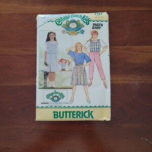 Puede incluir: Un sobre de patrón de costura Butterick Cabbage Patch Kids, número 3141, para tallas 7-8-10. El frente presenta ilustraciones de tres conjuntos: un vestido azul claro, una blusa azul con falda a cuadros y una blusa a cuadros con pantalones rosas.
