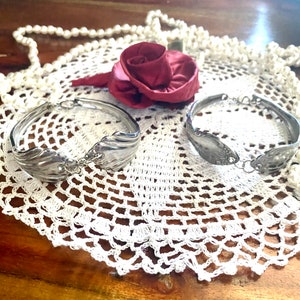May include: Two silver spoon bracelets on a white crocheted doily. The bracelets are made from bent silverware with a decorative design.