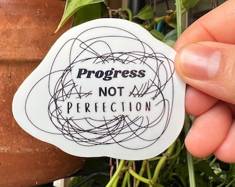 May include: A white die-cut sticker with an irregular shape featuring a black ink scribble design. The text Progress NOT PERFECTION is printed in the center in a combination of bold serif and clean sans-serif black lettering.