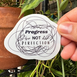 May include: A white die-cut sticker with an irregular shape featuring a black ink scribble design. The text Progress NOT PERFECTION is printed in the center in a combination of bold serif and clean sans-serif black lettering.