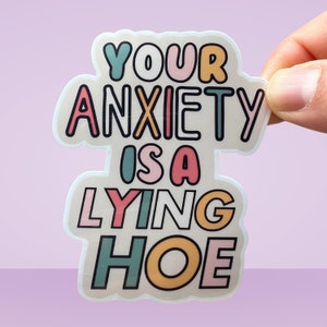 Może przedstawiać: Naklejka z napisem "YOUR ANXIETY IS A LYING HOE" w kolorowej, zabawnej czcionce. Naklejka ma przezroczystą obw&oacute;dkę i jest trzymana na fioletowym tle.