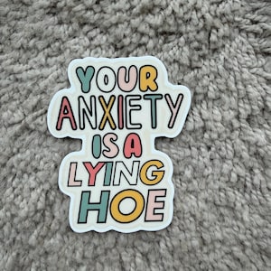 Może przedstawiać: Biała naklejka z napisem "YOUR ANXIETY IS A LYING HOE" w kolorowej, zabawnej czcionce. Tekst jest obrysowany na czarno i zawiera odcienie turkusu, r&oacute;żu, ż&oacute;łtego i pomarańczowego.