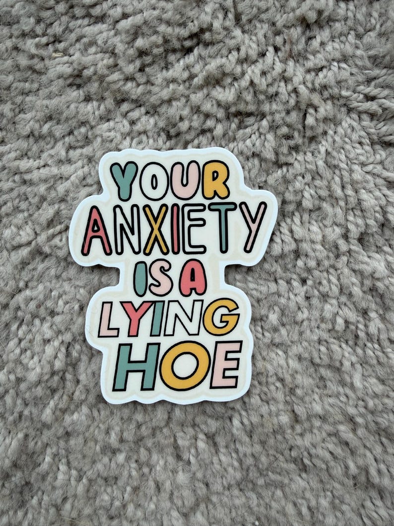 Może przedstawiać: Biała naklejka z napisem "YOUR ANXIETY IS A LYING HOE" w kolorowej, zabawnej czcionce. Tekst jest obrysowany na czarno i zawiera odcienie turkusu, r&oacute;żu, ż&oacute;łtego i pomarańczowego.