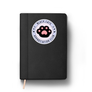 May include: Black notebook with a circular sticker featuring a black cat paw print with a pink pad. The sticker reads "Black Cats Appreciation Club" in a circular design with small heart accents. A light-colored ribbon bookmark is visible.