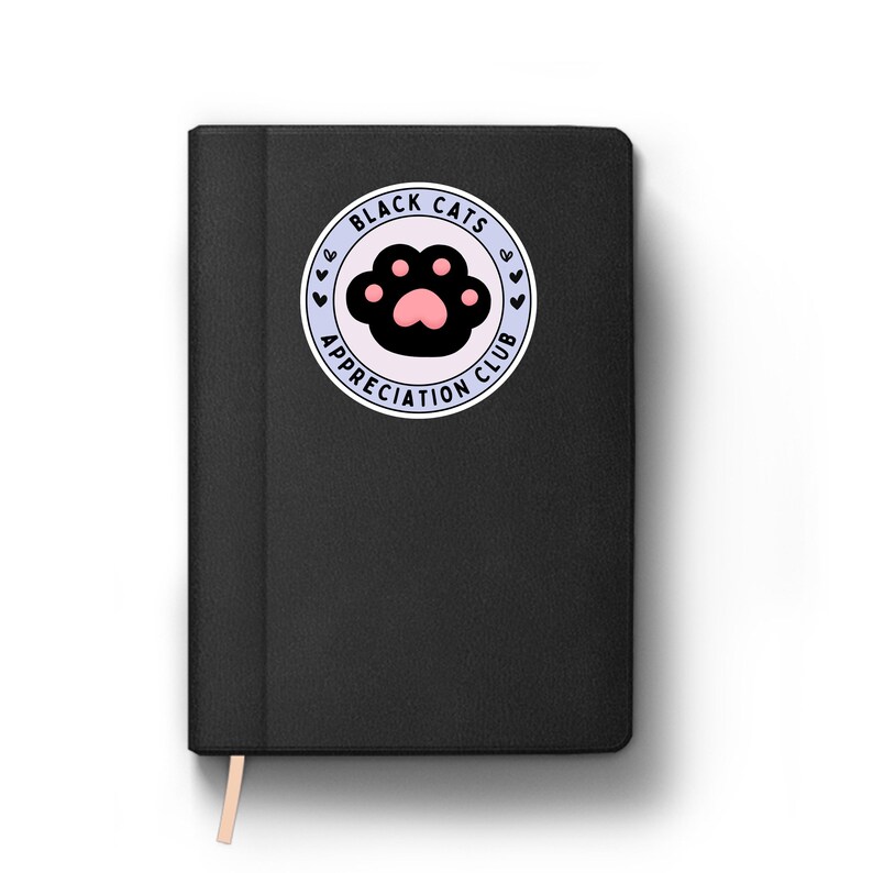 May include: Black notebook with a circular sticker featuring a black cat paw print with a pink pad. The sticker reads "Black Cats Appreciation Club" in a circular design with small heart accents. A light-colored ribbon bookmark is visible.