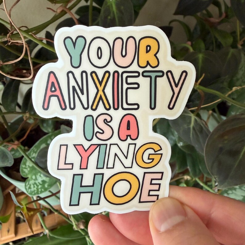 Może przedstawiać: Biała naklejka z napisem "YOUR ANXIETY IS A LYING HOE" w kolorowej, zabawnej czcionce. Naklejka ma czarny kontur i jest trzymana na tle zielonych roślin.