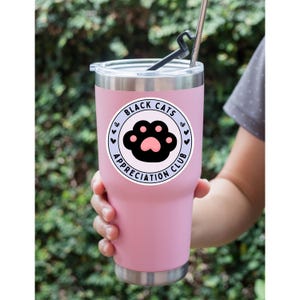 May include: A pink insulated tumbler with a silver rim and lid. The tumbler features a circular sticker with the text "Black Cats Appreciation Club" and a black cat paw print design. A reusable metal straw is inserted into the lid.