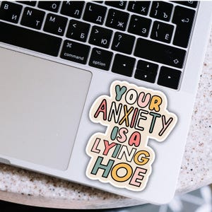 Może przedstawiać: Kolorowa naklejka z napisem "YOUR ANXIETY IS A LYING HOE" w zabawnej czcionce. Naklejka znajduje się na laptopie, z widoczną klawiaturą. Tekst jest w r&oacute;żnych kolorach, w tym r&oacute;żowym, turkusowym i ż&oacute;łtym.